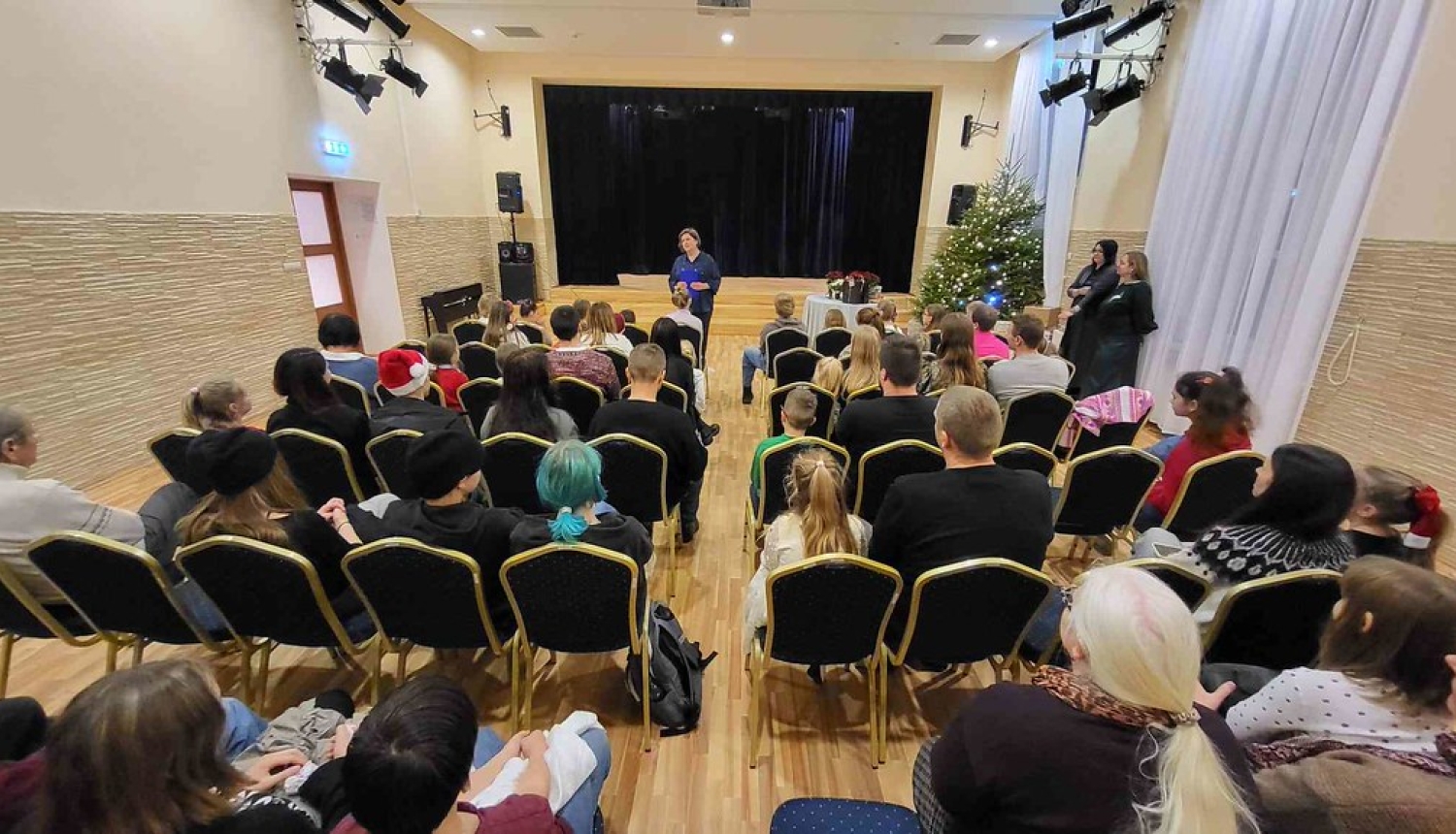 Tautas nama zālē Ķeguma audzēkņi sēž uz krēsliem un pedagogs runā priekšā uz skatuves